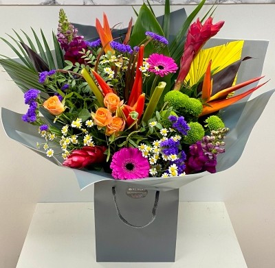 Vibrant Mother's Day bouquet featuring tropical flowers like ginger and heliconias alongside classic roses, germini, and daisy matricaria, wrapped in signature cellophane with a matching gift bag.