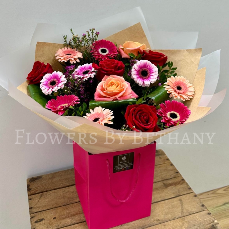 A beautifully arranged bouquet of red roses, mixed pink gerbera daisies, peach roses, and lush greenery, presented in a pink gift bag.