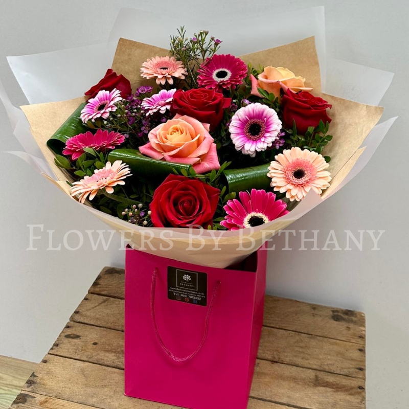 A beautifully arranged bouquet of red roses, mixed pink gerbera daisies, peach roses, and lush greenery, presented in a pink gift bag.