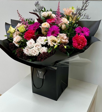 A beautiful bouquet containing roses, spray roses, germini, lizianthus, carnations, limonium, astilbe and skimmia in rich pink and nude tones, presented in a black gift bag and wrap. 