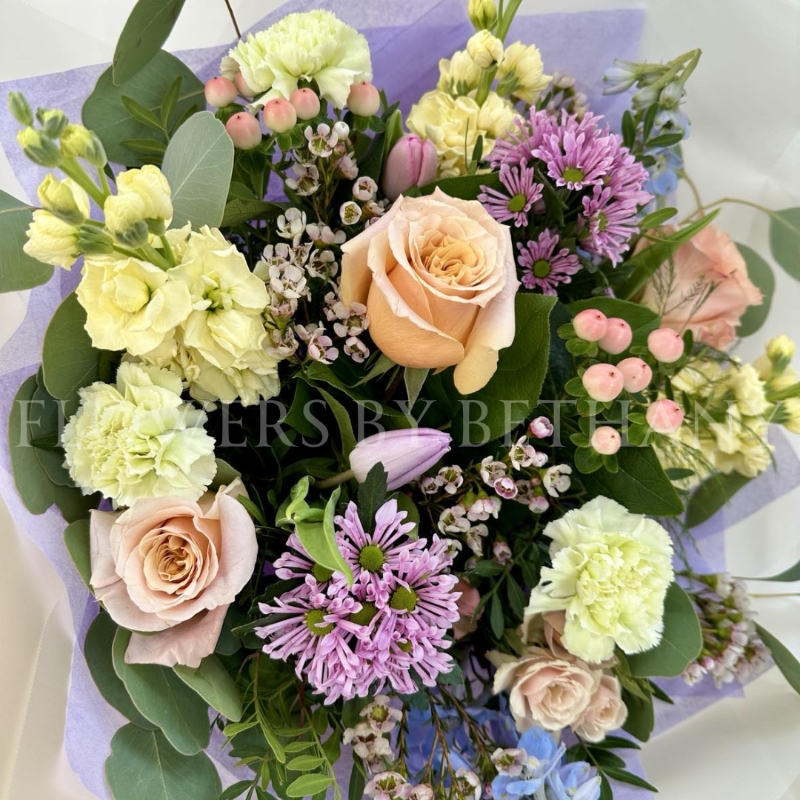 A pastel bouquet featuring peach roses, lilac tulips, pink hypericum, lemon stocks, and seasonal blooms, elegantly arranged with mixed foliage and presented in our signature gift bag with handles.