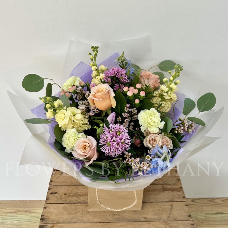 A pastel bouquet featuring peach roses, lilac tulips, pink hypericum, lemon stocks, and seasonal blooms, elegantly arranged with mixed foliage and presented in our signature gift bag with handles.