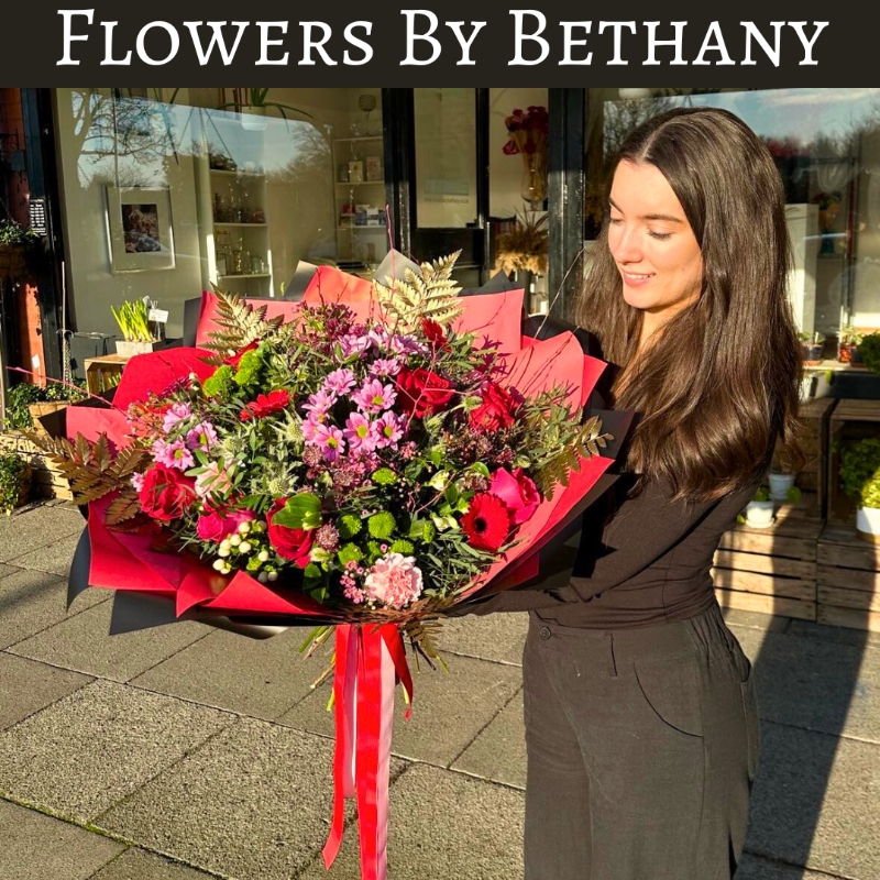 A luxurious bouquet featuring red roses, pink germini, green chrysanthemums, and gold-leaf accents among other seasonal florals. Wrapped in bold red and black wrapping with matching ribbon trails. 