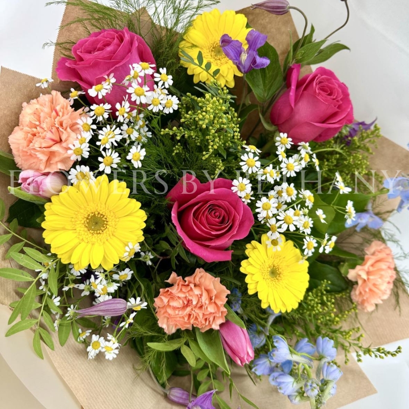 A vibrant, hand-tied bouquet featuring roses, daisies, delphinium, germini, carnations, and seasonal flowers, complemented with mixed foliage, wrapped in cellophane and presented in a gift bag with water.