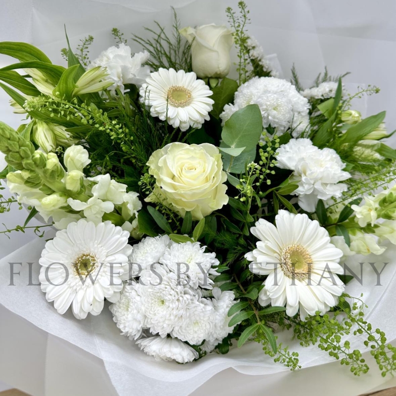 A serene, hand-tied bouquet featuring roses, carnations, alstroemeria, snapdragons, germini, and seasonal blooms, complemented with mixed foliage, wrapped in cellophane and presented in a gift bag with water.