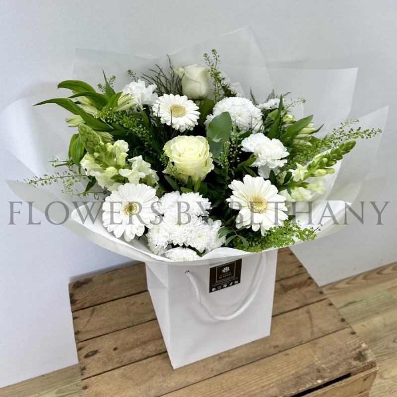 A serene, hand-tied bouquet featuring roses, carnations, alstroemeria, snapdragons, germini, and seasonal blooms, complemented with mixed foliage, wrapped in cellophane and presented in a gift bag with water.