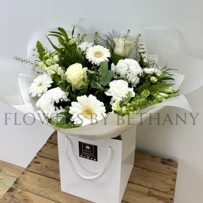 A serene, hand-tied bouquet featuring roses, carnations, alstroemeria, snapdragons, germini, and seasonal blooms, complemented with mixed foliage, wrapped in cellophane and presented in a gift bag with water.