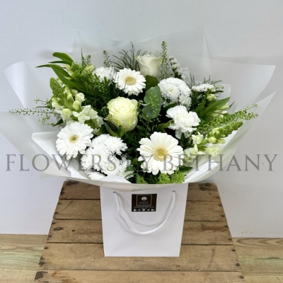 A serene, hand-tied bouquet featuring roses, carnations, alstroemeria, snapdragons, germini, and seasonal blooms, complemented with mixed foliage, wrapped in cellophane and presented in a gift bag with water.