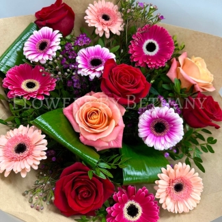 A beautifully arranged bouquet of red roses, mixed pink gerbera daisies, peach roses, and lush greenery, presented in a pink gift bag.