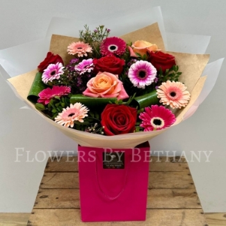 A beautifully arranged bouquet of red roses, mixed pink gerbera daisies, peach roses, and lush greenery, presented in a pink gift bag.