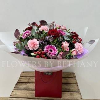 Berry Christmas bouquet featuring red roses, pink germini, carnations, spray roses, lilac limonium, and hypericum berries, wrapped in cellophane and presented in a burgundy gift bag.