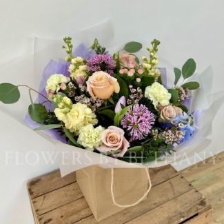 A pastel bouquet featuring peach roses, lilac tulips, pink hypericum, lemon stocks, and seasonal blooms, elegantly arranged with mixed foliage and presented in our signature gift bag with handles.