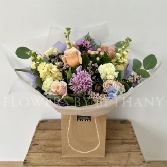 A pastel bouquet featuring peach roses, lilac tulips, pink hypericum, lemon stocks, and seasonal blooms, elegantly arranged with mixed foliage and presented in our signature gift bag with handles.