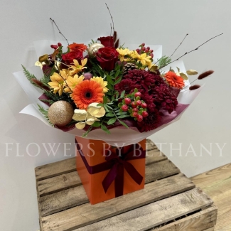 Fiery Festivities bouquet featuring orange germini, red chrysanthemums, red hypericum, red roses, pine cones, and gold baubles, presented in a burnt orange gift box with Christmas bow detailing