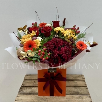 Fiery Festivities bouquet featuring orange germini, red chrysanthemums, red hypericum, red roses, pine cones, and gold baubles, presented in a burnt orange gift box with Christmas bow detailing