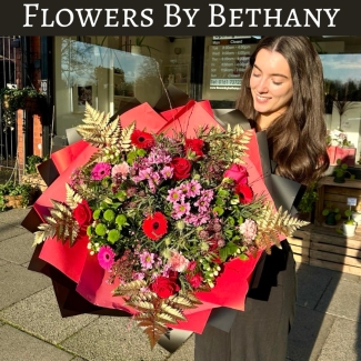 A luxurious bouquet featuring red roses, pink germini, green chrysanthemums, and gold-leaf accents among other seasonal florals. Wrapped in bold red and black wrapping with matching ribbon trails. 