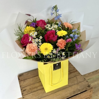 A vibrant, hand-tied bouquet featuring roses, daisies, delphinium, germini, carnations, and seasonal flowers, complemented with mixed foliage, wrapped in cellophane and presented in a gift bag with water.