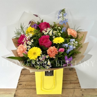 A vibrant, hand-tied bouquet featuring roses, daisies, delphinium, germini, carnations, and seasonal flowers, complemented with mixed foliage, wrapped in cellophane and presented in a gift bag with water.