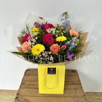 A vibrant, hand-tied bouquet featuring roses, daisies, delphinium, germini, carnations, and seasonal flowers, complemented with mixed foliage, wrapped in cellophane and presented in a gift bag with water.